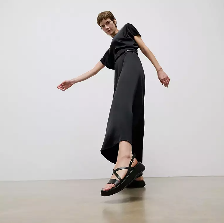 Black leather slingback crisscross sandal with low wedge and gold adjustable buckle on person.